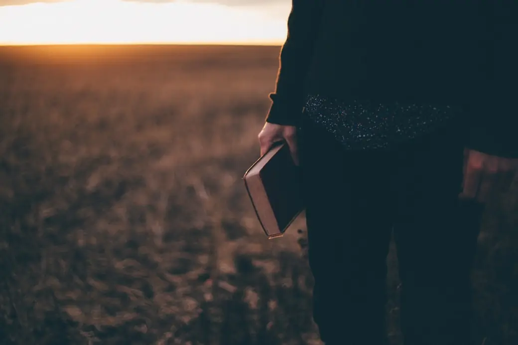A person holding a Bible while walking through a field at dusk
